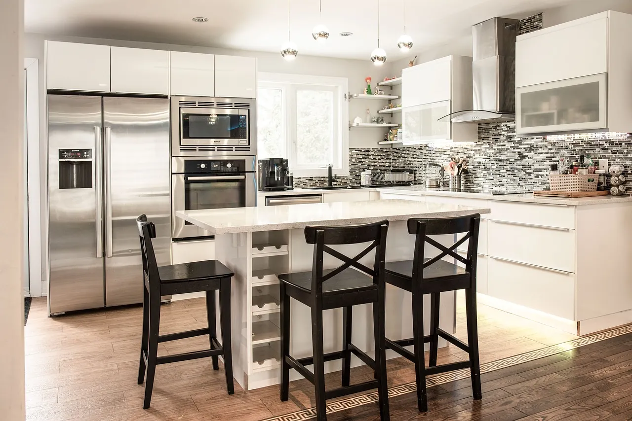 Modern kitchen with white cabinets, stainless steel appliances, a kitchen island with three black chairs, pendant lights, a tiled backsplash, and wooden flooring. The space is bright and organized.