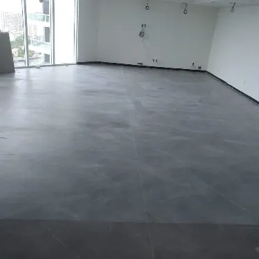 An empty room with a smooth, dark gray tiled floor, white walls, and large windows letting in natural light. Some electrical outlets and fixtures are visible on the far wall.