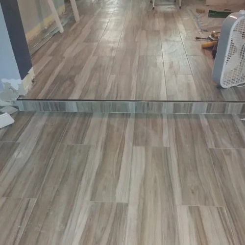 A room with wood-look tile flooring and a mirror on the wall, making it appear as though the floor continues seamlessly. Construction tools and a fan are visible in the background.