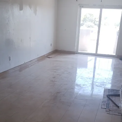 An empty room with shiny tiled floors, plain white walls, and a large glass sliding door letting in natural light. A small vent and a few tools or materials are visible on the floor near the door.