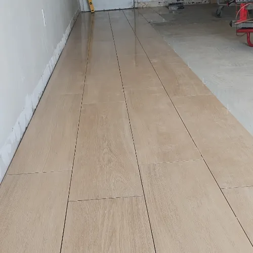 Light brown floor tiles with a wood-like pattern are installed in a row along a wall, partially completed, with construction materials visible in the background.