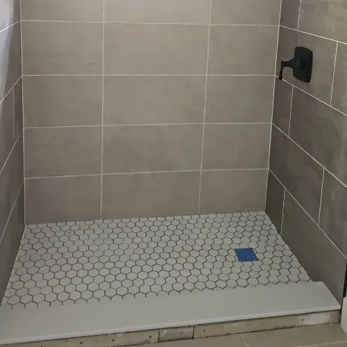 A tiled shower with beige wall tiles, a black showerhead and handle, and a white hexagonal tile floor. There is a small blue square on the floor and a white threshold at the entrance.