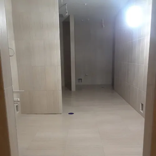 A mostly empty, unfinished room with light-colored tiled floors and walls, visible plumbing outlets, and a bright light source near the ceiling on the right side.