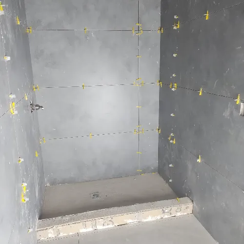 A shower area under construction with large grey tiles on the walls, yellow tile spacers visible between tiles, and a raised ledge at the entrance. No fixtures or shower fittings are installed yet.